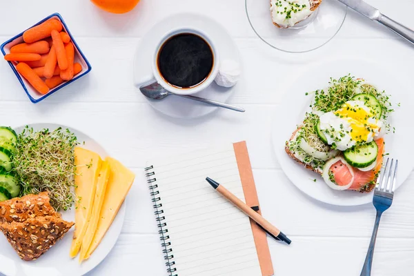 Top view planning notebook with copy space and healthy breakfast. Sandwich with egg benedict and smoked salmon, plate with cheese, sprout micro greens and cucumber, carrots and coffee on wooden table. — Stock Photo, Image
