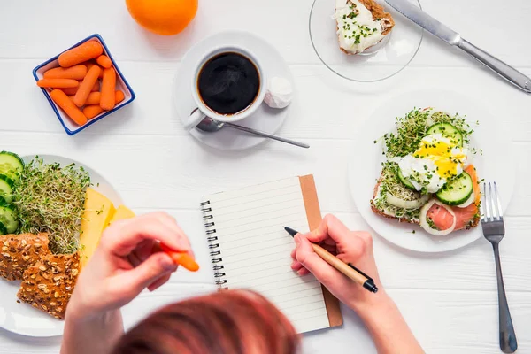 Morgengewohnheiten erfolgreicher Menschen. Tagesplanung und gesundes Essen. Frau isst Möhre und schreibt in Notizbuch auf den zum Frühstück servierten weißen Holztisch. Draufsicht, selektiver Fokus. — Stockfoto