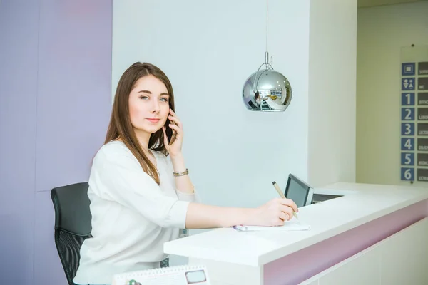 Portrét přátelské mladé ženy při pohledu na fotoaparát, mluví o telefonu za recepci. Správce v kanceláři, kliniky, centra, hotel. Okupace koncept. Selektivní fokus, prostor pro text. — Stock fotografie