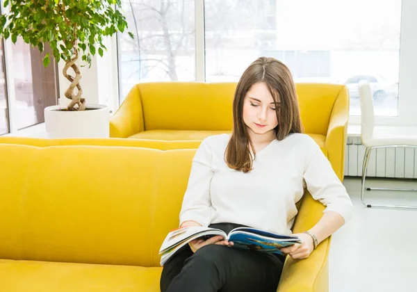 Portrait of young girl sitting on bright yellow sofa in modern waiting hall and reading a magazine. Hospitality, medicine, business concept. Selective focus. Space for text. — Stock Photo, Image