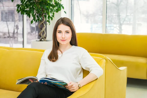 Ritratto di giovane ragazza che guarda la macchina fotografica, seduta su un divano giallo brillante in una moderna sala d'attesa e legge una rivista. Ospitalità, medicina, business concept. Concentrazione selettiva. Spazio per testo . — Foto Stock