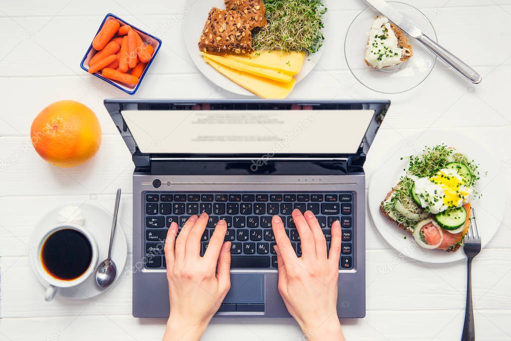 Top view female hands working on laptop and having healthy breakfast on served white wooden table with dishes. Working during eating. Day planning concept. Selective focus.