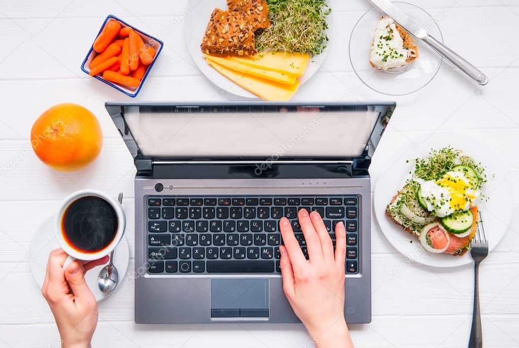 Top view female hands working on laptop and drink coffee on served white wooden table with healthy breakfast dishes. Working during eating. Day planning concept. Selective focus.