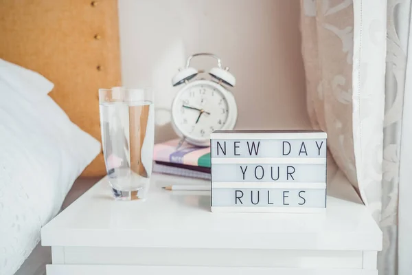 Good morning concept. Inspiration Motivational Life Quotes New day your rules message on lightened box with alarm clock, notebooks and glass of water on the bedside table in the sunlight. Copy space.