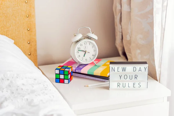 Good morning concept. Inspiration Motivational Life Quotes New day your rules message on lightened box with alarm clock, notebooks and rubik\'s cube on the bedside table in the sunlight. Copy space.