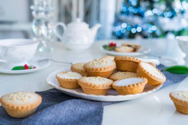 Geleneksel İngiliz usulü pasta kıymalı turtalar çay saatinde servis edilir ve arka planda ışıklandırılmış noel ağacı vardır. Sıcak bir ev havası. Yaklaş, seçici odaklanma. Boşluğu kopyala.