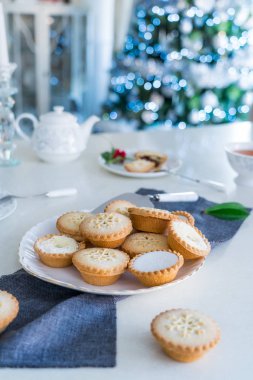 Geleneksel İngiliz usulü pasta kıymalı turtalar çay saatinde servis edilir ve arka planda ışıklandırılmış noel ağacı vardır. Sıcak bir ev havası. Dikey kart. Yaklaş, seçici odaklanma. Boşluğu kopyala.