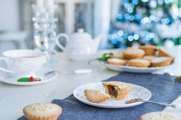 Tea time table setting with broken Mince Pie with filling on white plate. Traditional english festive pastry with lightened christmas tree on background. Cozy home mood. Close up, selective focus. — Stock Photo, Image