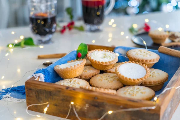 Nice warm cosy composition of traditional english festive pastry mince pies in wooden tray with mulled wine drinks and lights garland on home table with christmas tree on background. Close up. — Stock Photo, Image