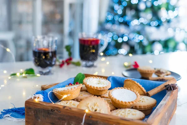 Nice warm cosy composition of traditional english festive pastry mince pies in wooden tray with mulled wine drinks and lights garland on home table with christmas tree on background. Close up. — Stock Photo, Image