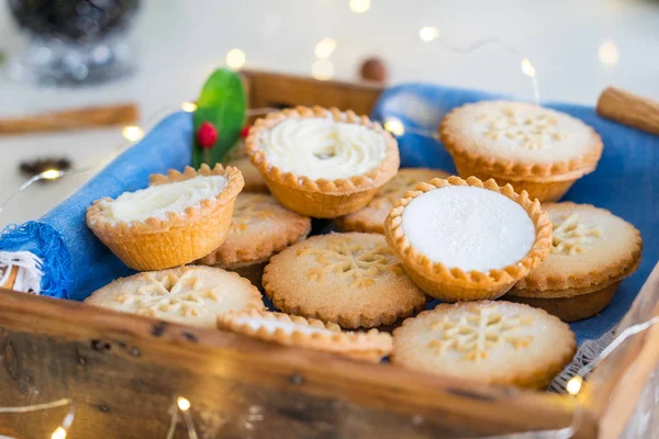 Close up traditional english festive pastry mince pies on wooden tray with blue napkin with blurred mulled wine drinks and lights garland on home table background. Soft selected focus. — Stock Photo, Image