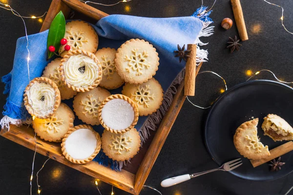 Top view warm cosy composition of traditional english festive pastry mince pies in wooden tray, spices, black plate with broken pie and lights garland on black background. Close up. Selective focus. — Stock Photo, Image