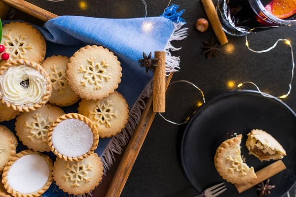 Top view warm cosy composition of traditional english festive pastry mince pies in wooden tray, spices, black plate with broken pie and lights garland on black background. Close up. Selective focus. — Stock Photo, Image