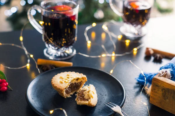 Close up broken Mince Pie with filling on black plate, mulled wine drinks and lights garland on dark table background. Traditional english festiveChristmas pastry. Soft selected focus. — Stock Photo, Image