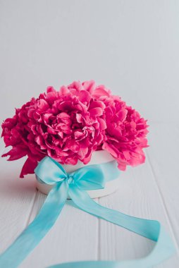 Turkuaz uydu kurdelesiyle beyaz ahşap arka planda duran yuvarlak kutuda güzel pembe şakayık çiçeği buketi. Hediye, selam, iltifat. Dikey kart. Yumuşak seçici odaklanma. Boşluğu kopyala