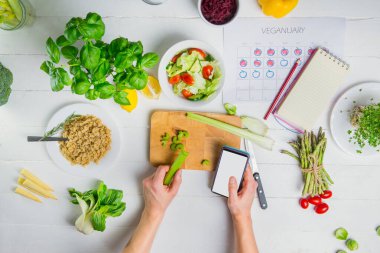 Üst Manzara Adamı 'nın elleri beyaz ekranlı akıllı telefonu ve kesme tahtasının üzerinde kereviz çubuğu tutuyor. Sebzeli taze vegan salatası pişiriyorum. Vejetaryen takvimi ve günlük diyet planlaması