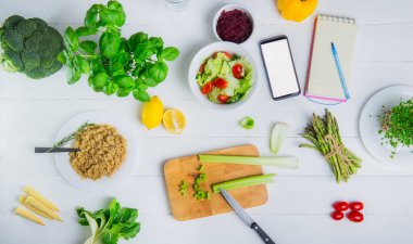 Her gün sağlıklı beslenme planlaması. Çiğ sebze, taze salata, defter ve telefon beyaz ahşap masada. Vejetaryen ve vejetaryen diyeti. Vejetaryenlik kavramı. Sürdürülebilir yaşam tarzı, bitkisel gıdalar