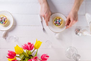 Üst düzey erkek eller, tabaklarda renkli tatlı yumurtalarla Paskalya masası servis ediyor. Beyaz ahşap masada kristal cam eşyalar ve taze lale çiçekleri. Aile tatili. Boşluğu kopyala