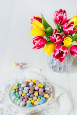 Bayramlık Paskalya şekerleri tabakta çeşitli pastel renklerde yumurtalar ve ahşap masa üzerinde taze laleler bulunan vazonun yanında kaliteli peçete ile kaplıydı. Aile tatili geçmişi. Aydınlık dikey kart. Boşluğu kopyala