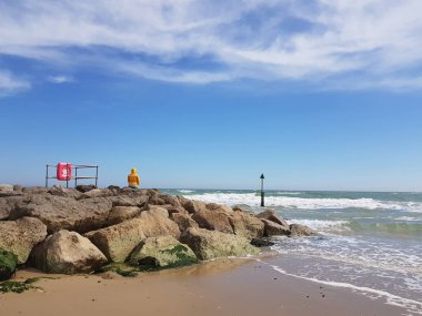 Parlak sarı ceketli kadın taş iskelede tek başına oturuyor ve güneşli bir günde deniz manzarası seyrediyor. Sosyal uzaklık kavramı. Telefon fotoğrafı. Boşluğu kopyala