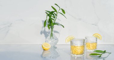 Limonla süslenmiş sarı kokteylli iki kristal bardak. Taze yeşillik vazosu ve yeşil çay kadehleri mermer zemin üzerinde alkol içeceği. Doğrudan güneş ışığı ve gölgeler. Minimalizm