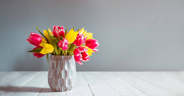 Fresh spring yellow and pink tulips bouquet in blue vase standing on black wooden table with gray background. Festive flowers for mother's or women's day. Mockup for greeting wide banner. Copy space