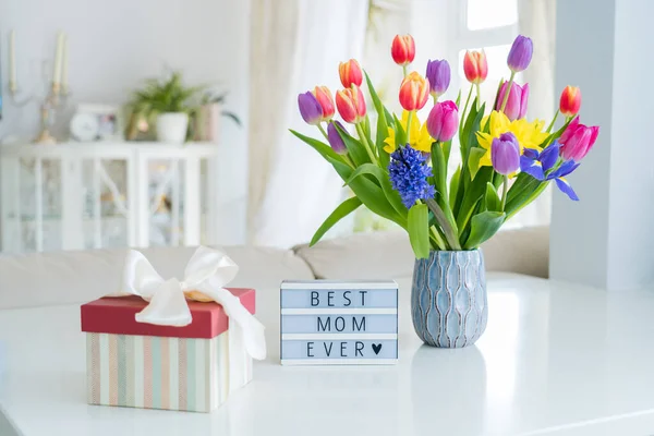 Happy Mother\'s Day background. Colorful spring flowers bouquet in vase, gift box with satin ribbon and lightbox with words Best mom ever on white marble table with light interior room view. Copy space.
