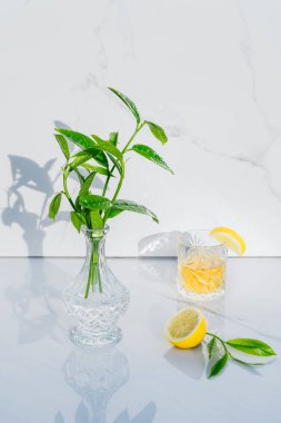 Limonun yarısı, kristal bardak, sarı kokteyl ve mermer zemin üzerinde taze yeşillik olan vazo. Yeşil çay ve alkol dolu kadehler. Doğrudan güneş ışığı ve gölgeler. Minimalizm. Boşluğu kopyala