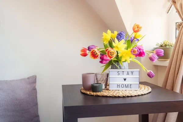 Everything for a calm stay at home. Lightbox with Stay home message, bouquet of fresh spring flowers and candles on the coffee table, and pillow sitting place. Cozy area for relaxing near window