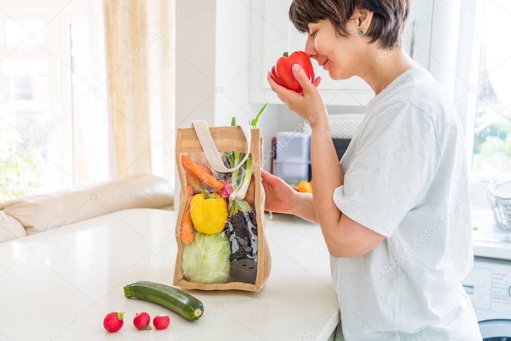 Woman taking and checking fresh organic vegetables from reusable eco sackcloth fabric bag packaging on white kitchen table. Home delivery concept. Local farmer healthy food. Zero waste. Copy space