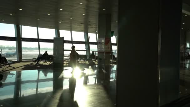 Airport terminal interior, people travellers passengers silhouettes waiting in a lounge, hall view, beautiful background with sunshine, dolly shot from escalator — Stock Video