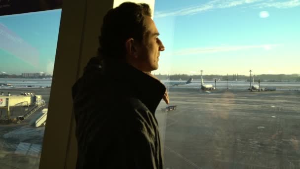 Hombre de pie en la ventana en el interior de la terminal del aeropuerto, personas pasajeros siluetas esperando en un salón, vista al pasillo, hermoso fondo con sol, dolly shot de escaleras mecánicas, viajes — Vídeos de Stock