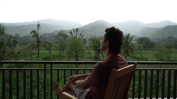 Mann entspannt auf Balkon mit wunderschönem Bergblick im Urlaub auf der Hotelterrasse, gut aussehender, sportlicher Mann in kurzen Shorts genießt schönen nebligen Morgenblick. — Stockvideo