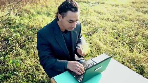 Молодий привабливий бізнесмен у костюмі з портативним комп'ютером, що сидить на зеленому лузі зовні, офіс — стокове відео