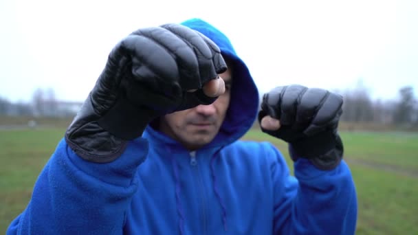 Человек спортсмен боксер парень в боксерских перчатках тренировки на открытом воздухе, делает удары в, человек в капюшоне — стоковое видео