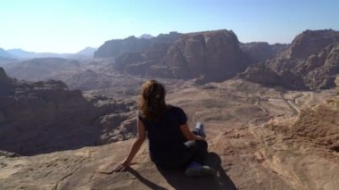 Kıvrımlı kızıl saçlı bir kadın rüzgarda esiyor dağın kenarında oturuyor antik harabelerin altında