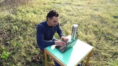 Bahçede uzaktan kumandalı çalışan genç bir müteahhit dizüstü bilgisayar ve masayla parkta oturuyor.