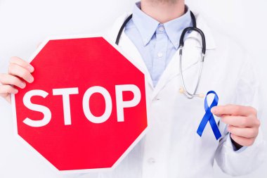 Onkolojik hastalık konsepti. Beyaz önlüklü ve steteskoplu doktor bağırsak kanserinin sembolü olarak koyu mavi kurdele ve dur işareti taşıyor..