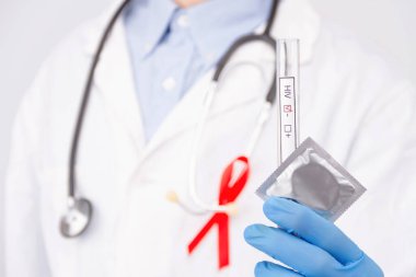 Safe sex / healthcare concept. Doctor wearing white coat with pinned red ribbon as a symbol of hiv aids and stethoscope holding negative tumor marker / blood test and condom.  clipart