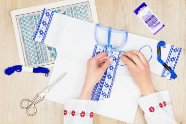 Geleneksel Ukrayna tişörtlü kız / kadın elleri çocuklar için nakış mavisi gömlek dikiyor. Arkaplanda nakış şeması, makas ve renkli iplikler.