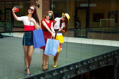 Renkli elbiseler, şapkalar ve yüksek topuklu ayakkabılar içinde alışveriş merkezinde alışveriş yaptıktan sonra selfie çeken oldukça mutlu, zeki bayan kızlar. Mağaza satışları, Kara Cuma.