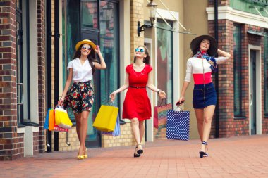 Renkli elbiseler, şapkalar ve yüksek topuklu ayakkabılar içinde alışveriş merkezlerinde alışveriş yaptıktan sonra sokakta yürüyen oldukça mutlu, zeki bayan kızlar. Mağaza satışları, Kara Cuma.