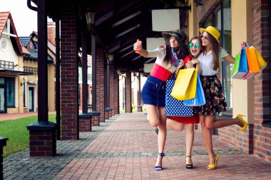 Renkli elbiseler, şapkalar ve yüksek topuklu ayakkabılar içinde alışveriş merkezinde alışveriş yaptıktan sonra selfie çeken oldukça mutlu, zeki bayan kızlar. Mağaza satışları, Kara Cuma.