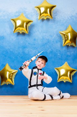 Astronot erkek çocuk, beyaz astronot kostümü içinde roketle astronotluk yapıyor ve yıldızların arasından evrene uçtuğunu hayal ediyor. Mavi arka planda altın yıldızın balonlarının yanında kalıyor.