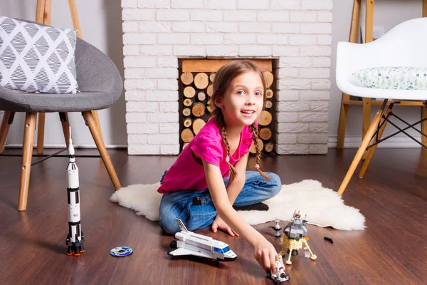 Young child girl female playing with cosmos\'s toys constructor: rocket, shuttle, rover, satellite and astronaut doll in comfortable interior at home on wooden floor