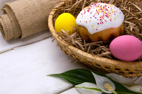 Easter cake and colored easter eggs in basket nest, sackcloth and flower on white wooden background
