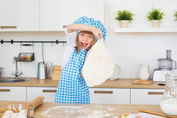 La bella bambina in vestiti di cuoco tiene in mani un cerchio di pasta su una cucina — Foto Stock