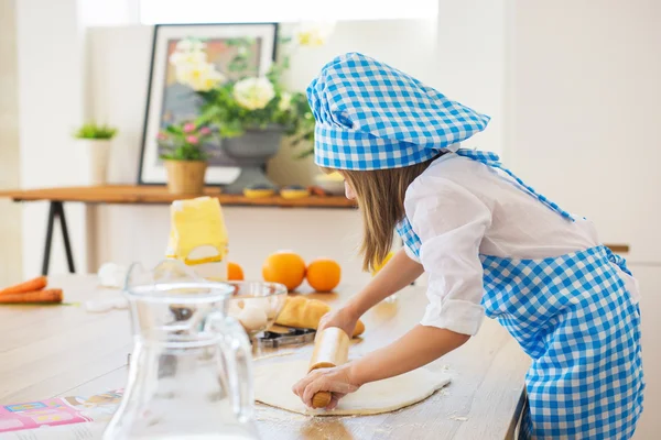 La bambina in vestiti di cuoco fa rotolare un cerchio di pasta su un tavolo — Foto Stock