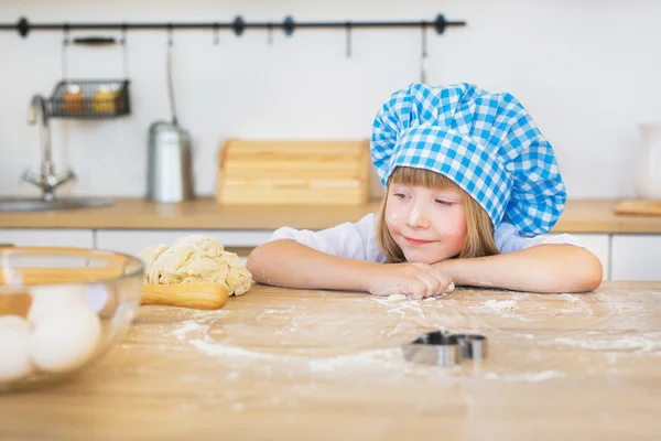 La bella bambina in un cuoco guarda su una pasta su un tavolo — Foto Stock