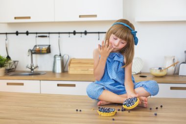 Sevimli küçük kız mavi elbiseli bir pasta yiyor ve bir tablo üzerinde parmaklarını yalıyor